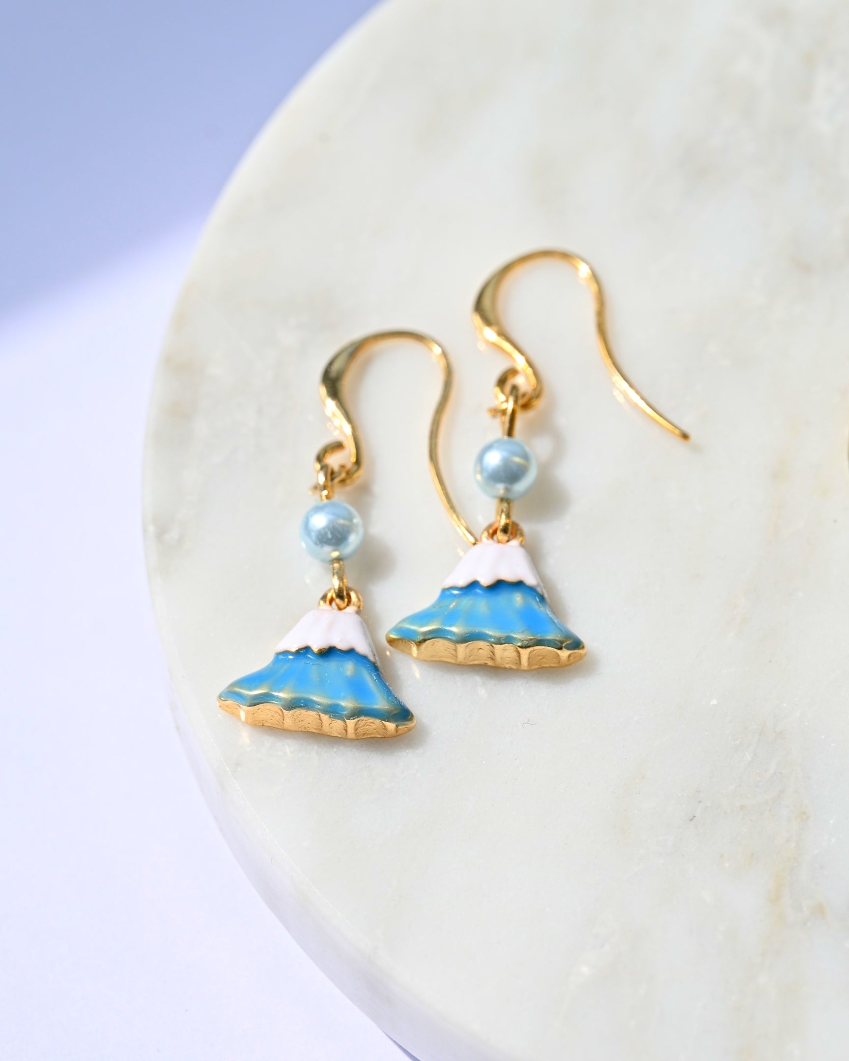 Pair of gold earrings with Mt Fuji charm and blue pearl on a marble background