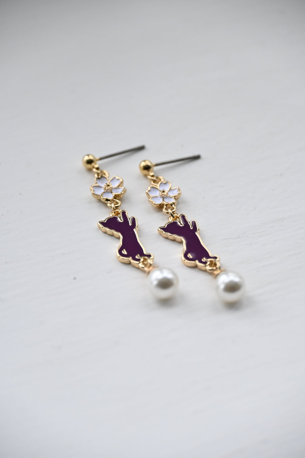 A close up shot of a pair of earring with white sakura and purple cat charms with white pearl on a white surface