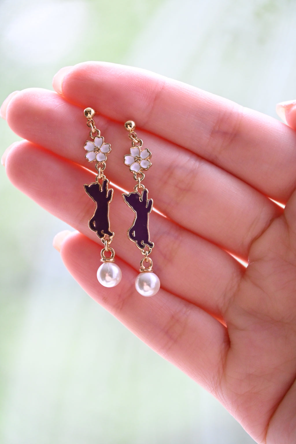 A pair of earring with white sakura and purple cat charms with white pearl against a hand