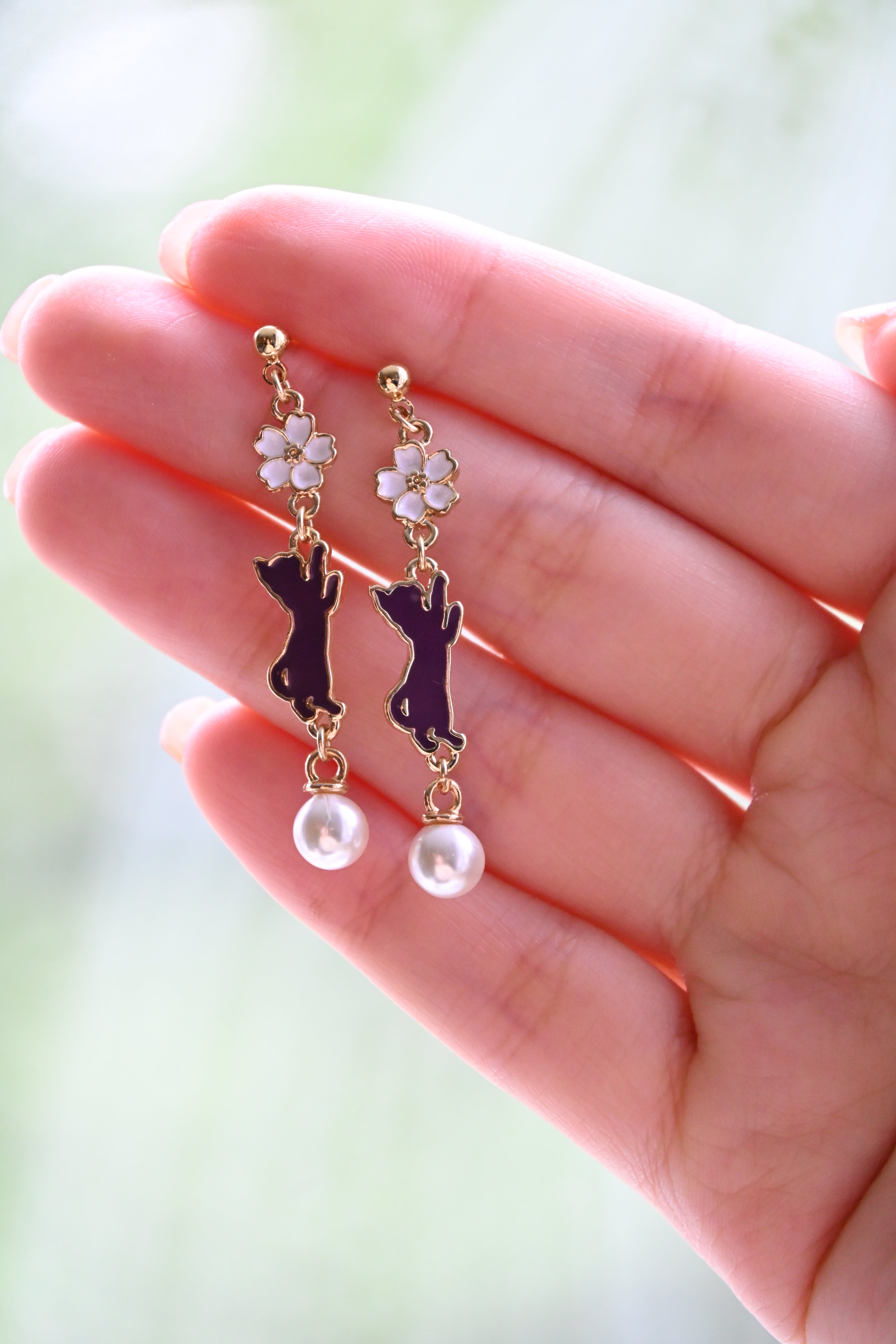 A pair of earring with white sakura and purple cat charms with white pearl against a hand