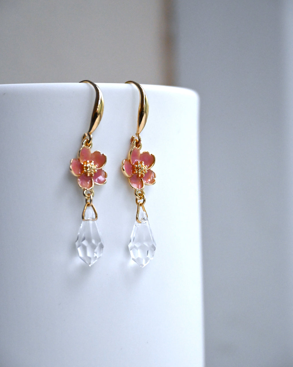 Gold earrings with pink sakura charms and clear teardrop beads on a white wall.
