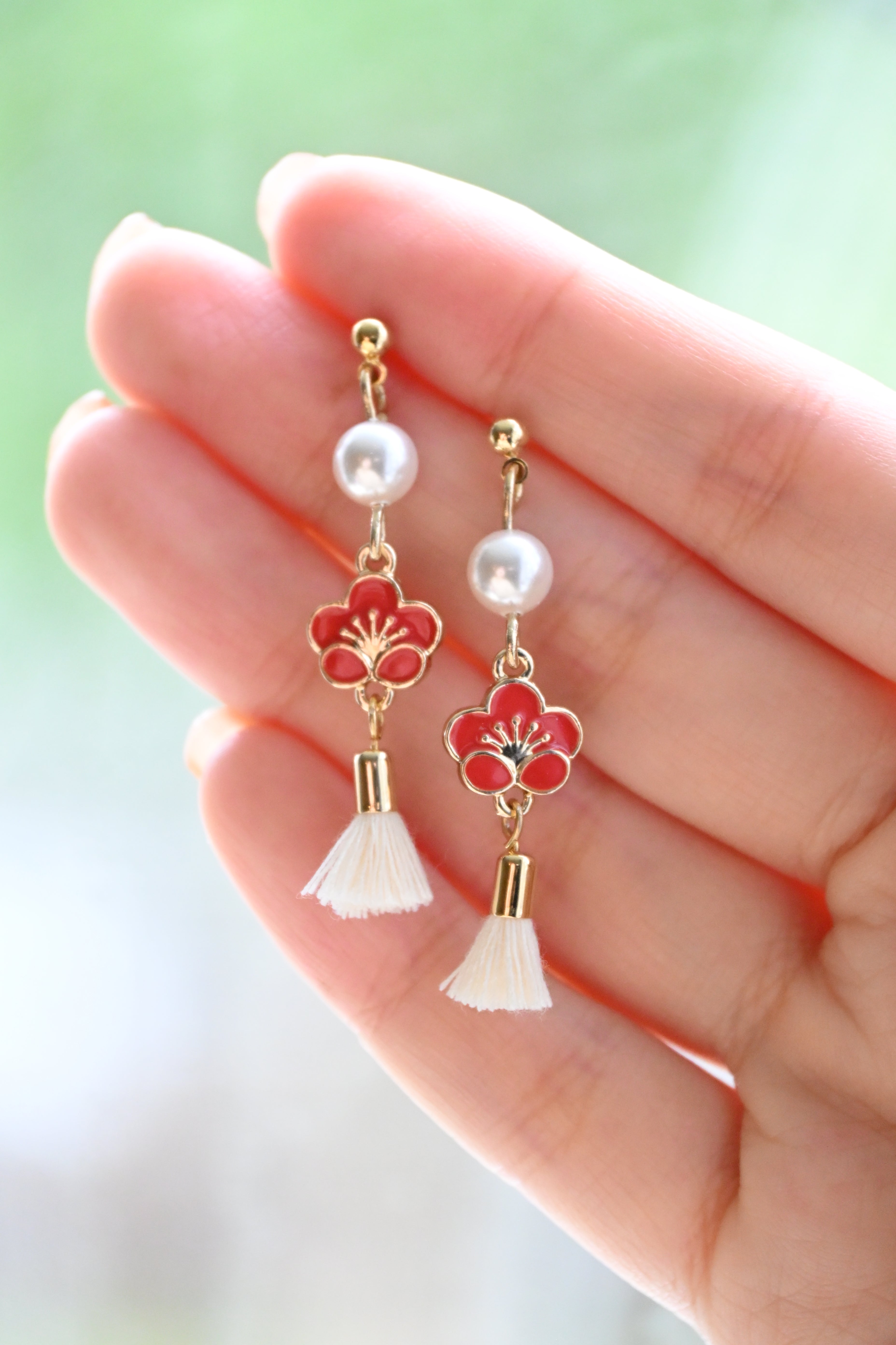 Earring with red plum flower charm with white pearl and tussel on a hand against a blurred background